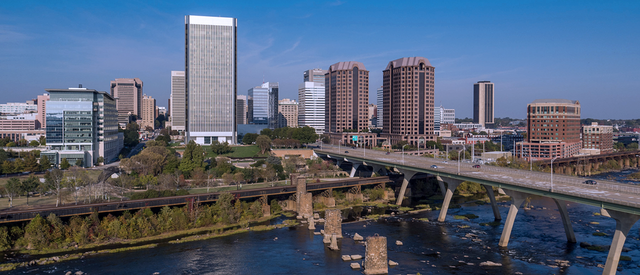 A city skyline with tall office buildings, a river in the foreground, and a highway bridge crossing over the water under a clear blue sky.